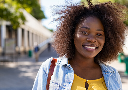 A young woman wearing a yellow blouse and blue denim jacket smiles.