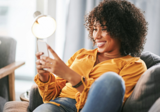 A woman wearing jeans and an orange blouse sits on a couch and scrolls on her phone.
