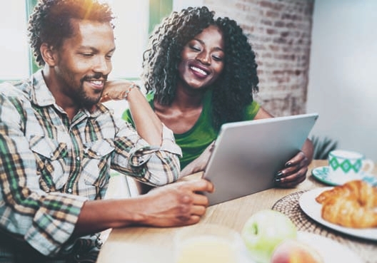 A couple sit at a table and enjoy croissants and tea while browsing an online magazine on a tablet.