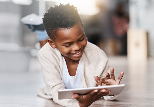 A little boy wearing a cream colored jersey lays on the floor and pays on a tablet.